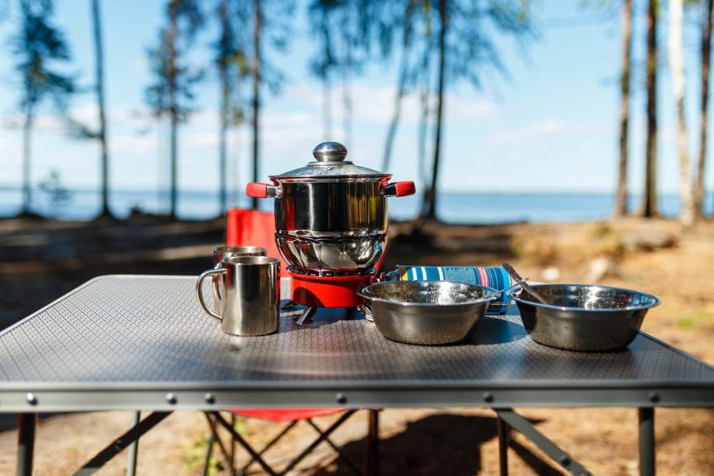 Cooking pans on fire from a portable gas burner next to plates and mugs on a camping table