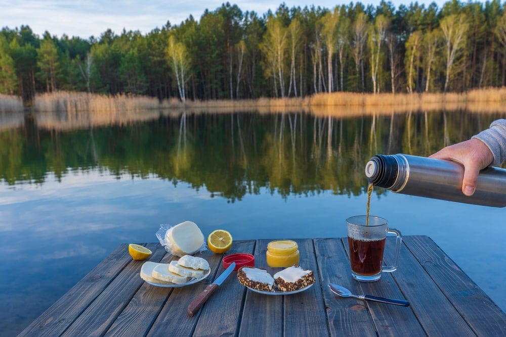 A person pouring coffee to a glass mug next to a camping breakfast on a wooden table