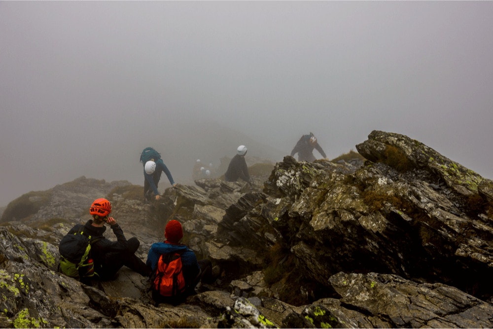 A Group of mountaineers wearing helmets take a rest on rocks on the top of the mountain while others are scrambling